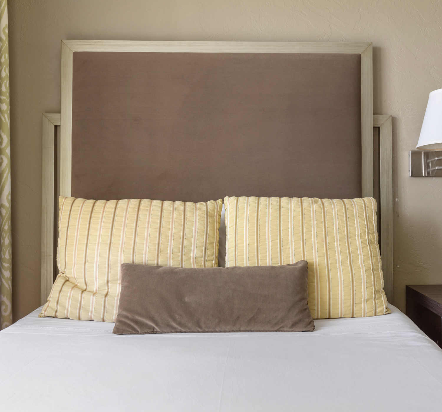 A neatly made bed with a brown cushioned headboard, two yellow pillows, a smaller brown pillow, a bedside table, and a white lamp.