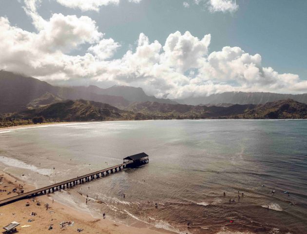 A scenic beach with people, a sandy shoreline, clear waters, a long pier extending into the ocean, and mountains in the background under a partly cloudy sky.
