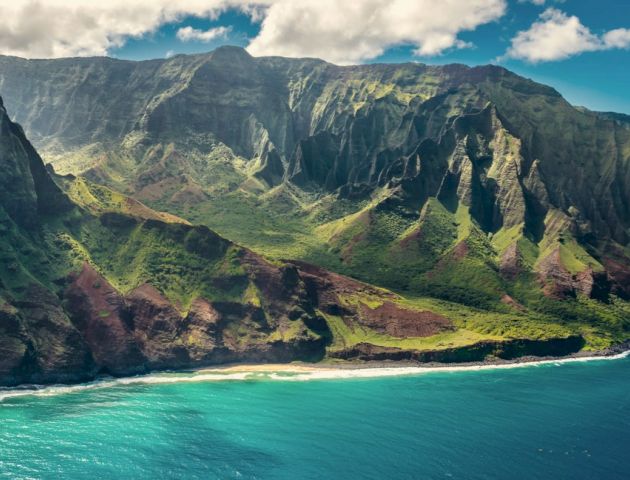 This image features a stunning coastal landscape with rugged, lush green mountains and clear, turquoise waters under a blue sky with scattered clouds.