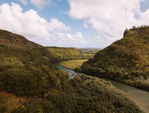 A scenic landscape features a meandering river surrounded by lush green hills under a partly cloudy sky, showcasing natural beauty.