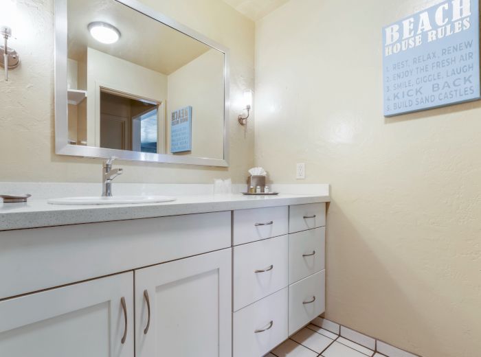 A bathroom with a large mirror, white cabinets, a "Beach House Rules" sign, a sink, and a light fixture is shown.