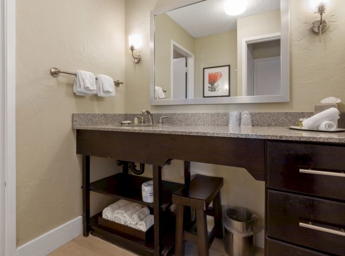 A bathroom with a large mirror, granite countertop, sink, open shelving with towels, wall lights, towel rail, and doorways to other rooms can be seen.