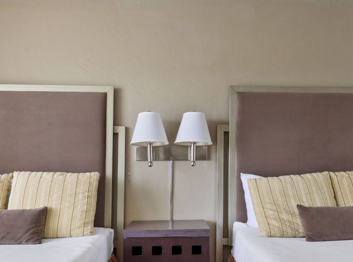 The image shows two identical beds with brown headboards, each with pillows. A small table with two lamps is placed between them with a beige wall behind.