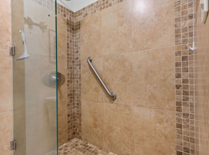A tiled shower with a glass door, grab bar, square tiles, and a built-in shelf in the corner.