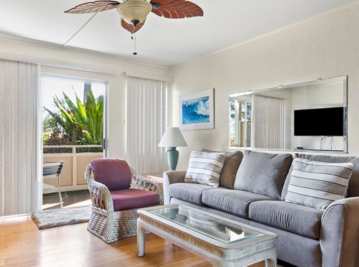 A bright living room with a gray sofa, striped pillows, a glass coffee table, wicker chair, ceiling fan, and a balcony with green plants is shown.