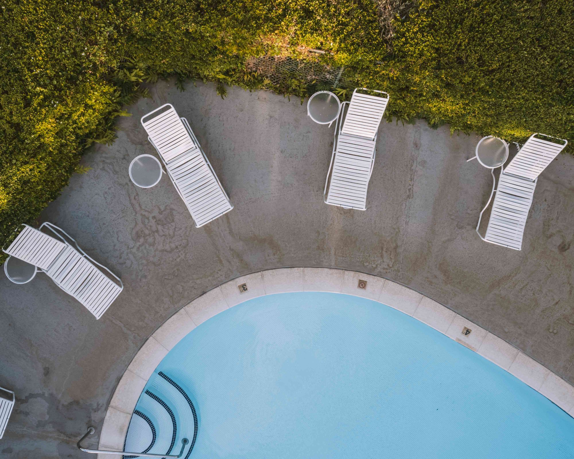 The image shows an aerial view of a swimming pool with white lounge chairs and tables surrounding it, situated near a grassy area.