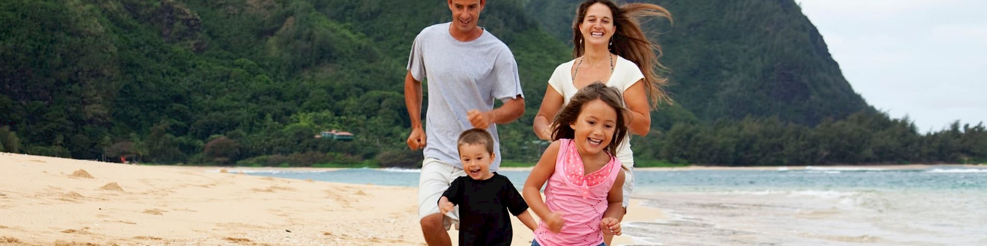 A family of four is happily running on a sandy beach, surrounded by lush green mountains and a calm sea in the background.