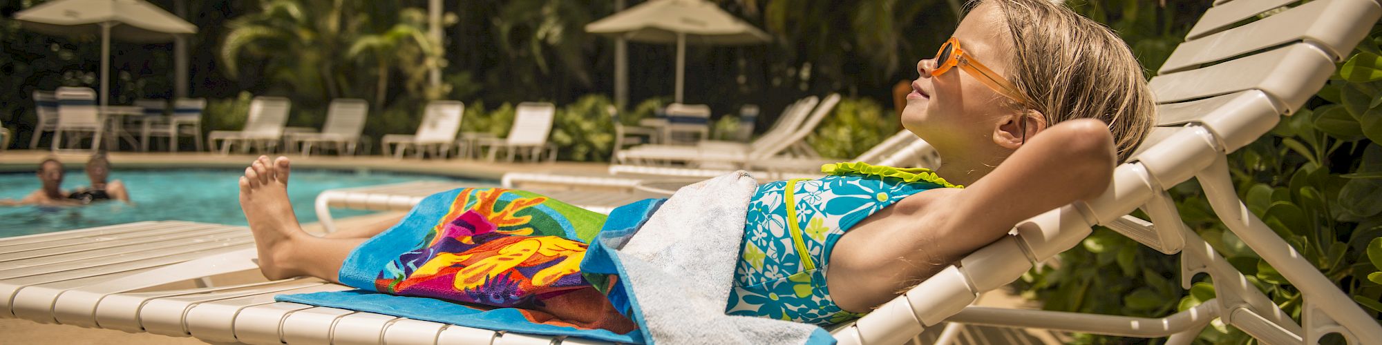 A child relaxes on a lounge chair by a pool, wearing sunglasses and wrapped in a colorful towel, surrounded by greenery and umbrellas.