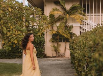 A woman in a flowing dress walks on a path beside a hedge, with a house and palm trees in the background.