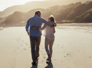 A couple walking arm in arm on a sandy beach with hills in the background, enjoying a peaceful and intimate moment together.