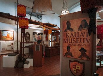 The image shows the interior of Kaua'i Museum with various exhibits, including a large canoe, historical artifacts, and informational panels.