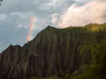 A scenic mountain landscape with steep cliffs covered in greenery, clouds overhead, and a faint rainbow appearing in the sky.