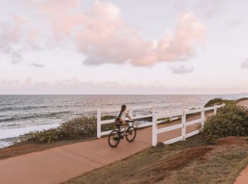 A person rides a bicycle on a paved path with a white fence, near the ocean, under a sky dotted with clouds.