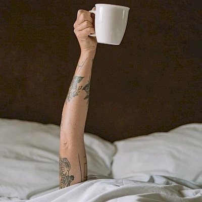 A tattooed arm is extending from under a blanket, holding a white coffee mug in the air.