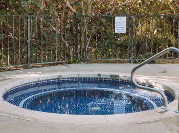 A round outdoor Jacuzzi is surrounded by a metal fence, with a handrail for easy access and a sign in the background, ending the sentence.