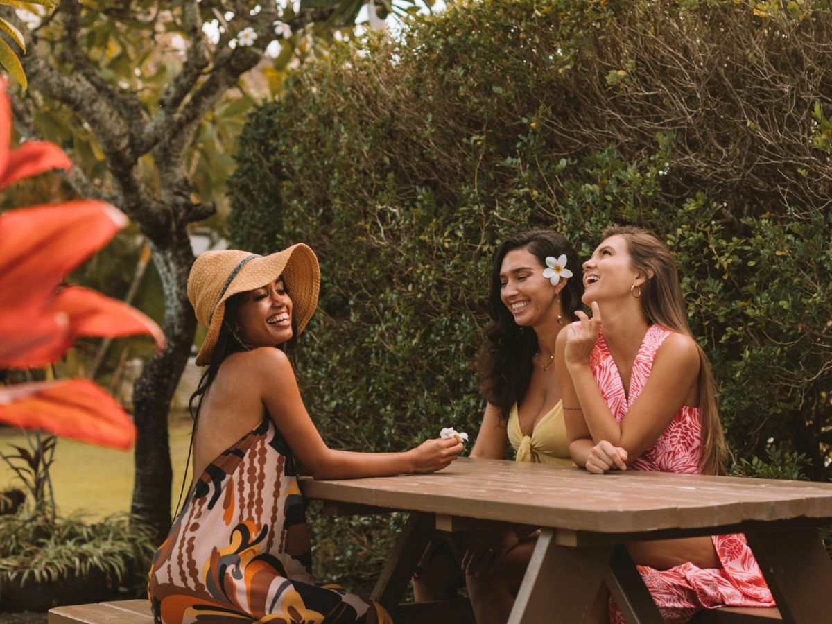Three women sit at a wooden picnic table in a garden, surrounded by greenery and trees, while engaged in conversation and smiling.