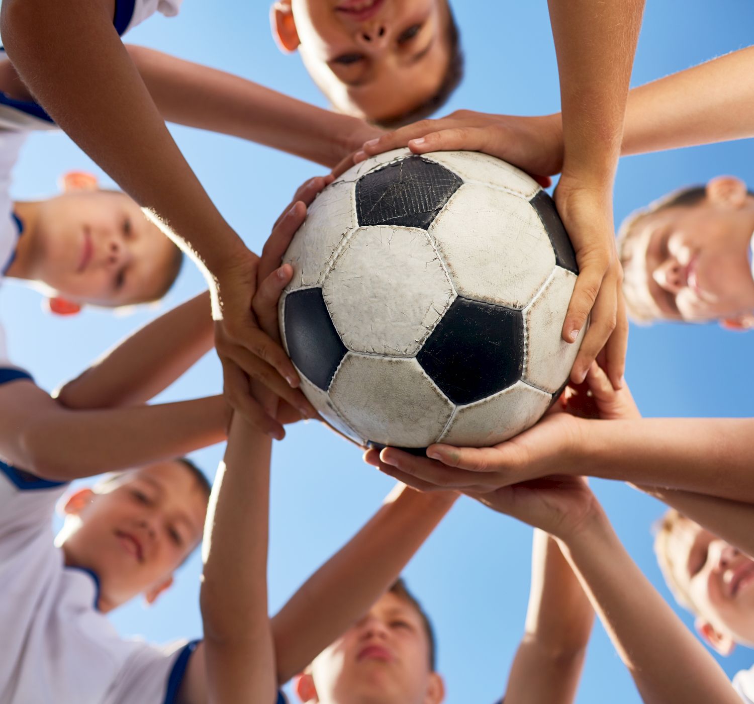 A group of boys in white shirts form a circle, each with one hand holding a soccer ball in the center, viewed from below against a blue sky.