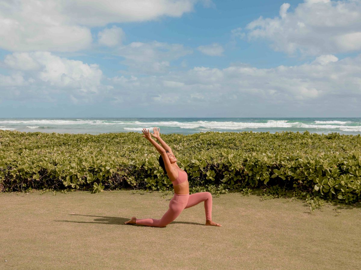 A person is performing a yoga pose on a grassy area near a beach with a clear blue sky and green shrubbery in the background.