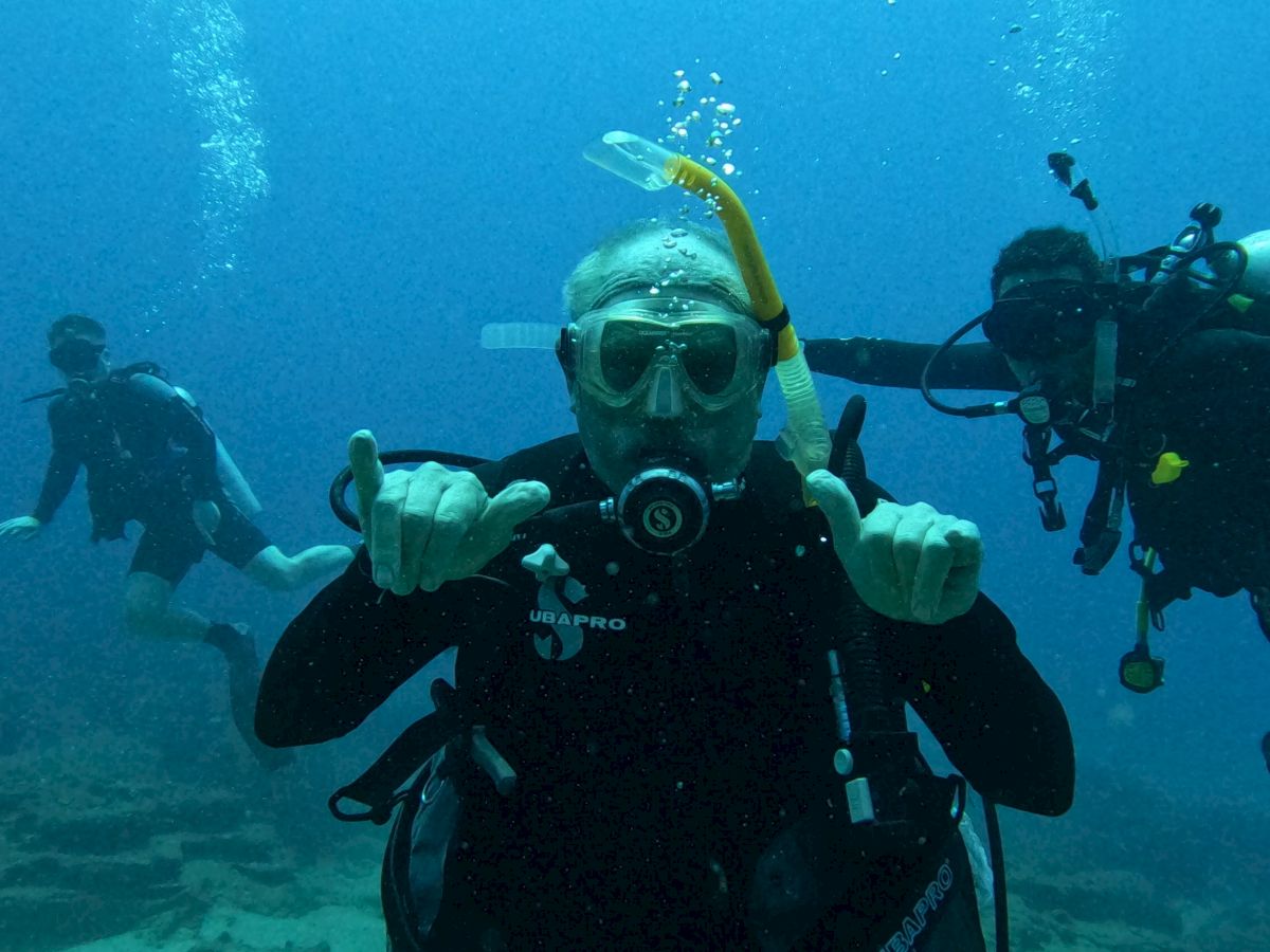 Three scuba divers are underwater, making hand signals and wearing diving gear. The background shows underwater bubbles and the ocean floor.