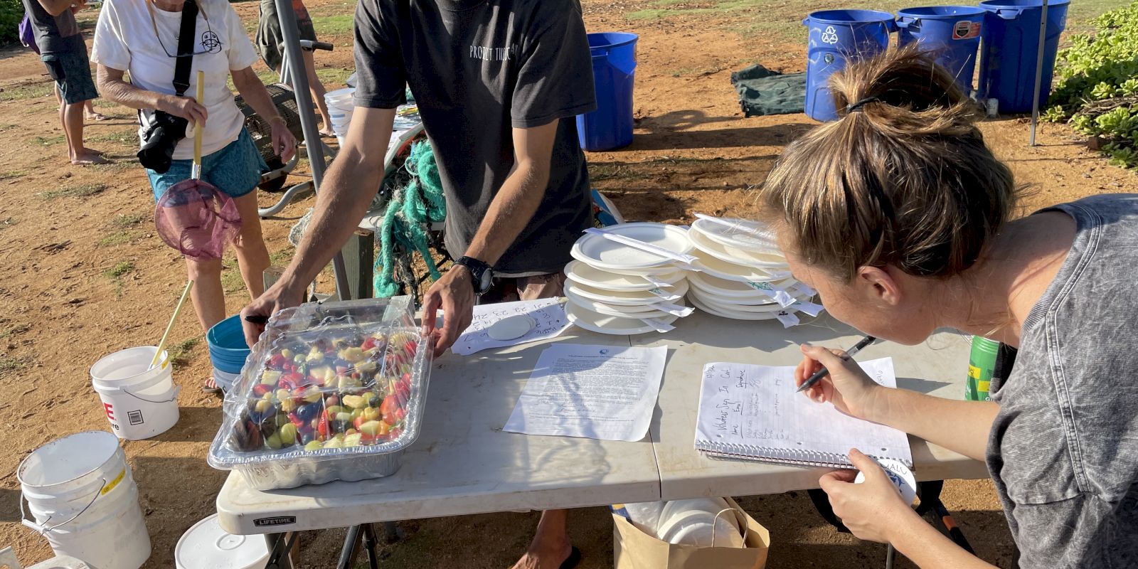 Two people work at an outdoor event table with trays, plates, and a clipboard. Buckets and blue bins are in the background. The sentence ends.