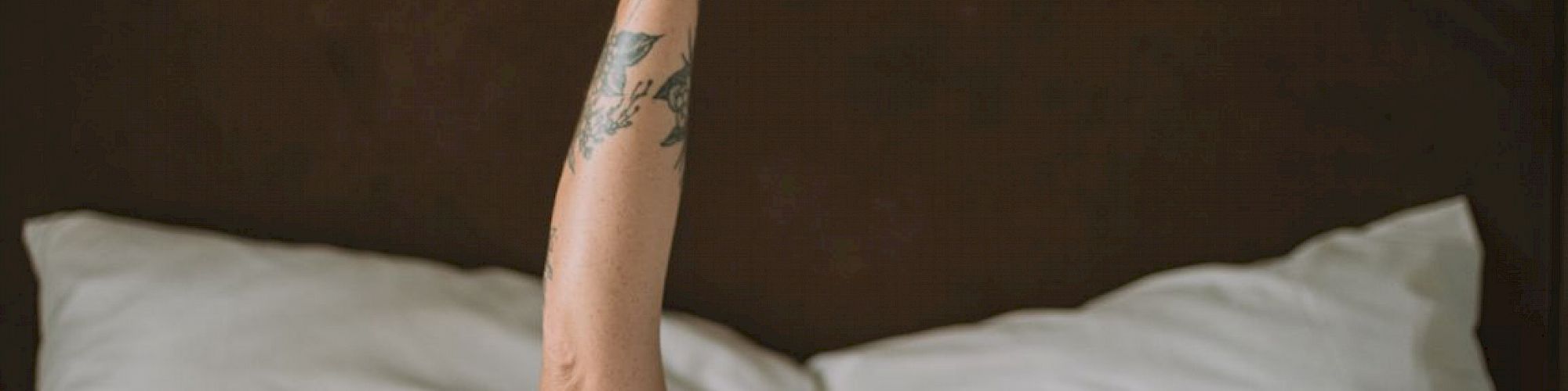 An arm with tattoos extends from beneath white bed covers, holding a white mug against a dark background.
